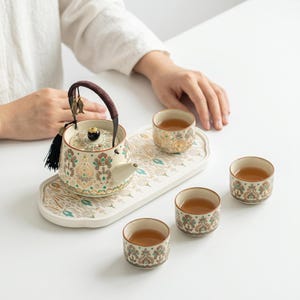 May include: A ceramic tea set featuring a teapot with a dark brown wrapped handle, a black tassel, and two small metal fish charms. The teapot and three matching cups are decorated with a repeating floral pattern in teal, brown, and cream.