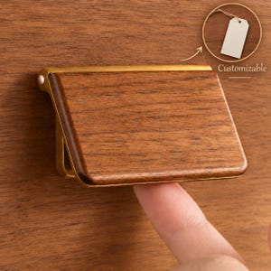 Puede incluir: Un tirador de mueble rectangular con un panel frontal de madera de grano marrón oscuro enmarcado por un borde de metal dorado cepillado. El herraje está montado sobre una superficie de madera a juego, con una etiqueta blanca que dice Customizable.