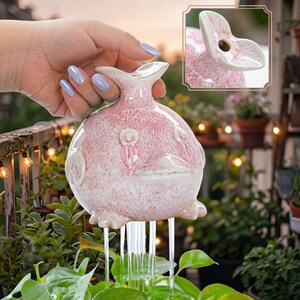 Pode incluir: Um vaso de cerâmica redondo, rosa e salpicado, em forma de pássaro, com uma abertura larga no topo. O pássaro apresenta um bico plano, pequenas asas e dois pés. Fluxos de água descem da base do vaso para a vegetação abaixo.