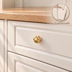 May include: A gold-toned metal cabinet knob shaped like a paw print, featuring a smooth, rounded finish. The knob is mounted on a white drawer front. A circular inset shows a blank white tag attached by a string with the text Customizable below.