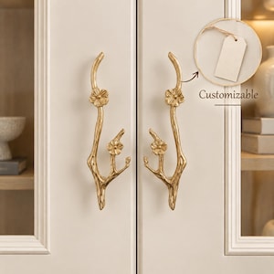 May include: Two gold-coloured metal cabinet handles shaped like tree branches with small flower blossoms. A circular inset shows a blank white rectangular tag hanging from a string with the text Customizable printed below the tag.