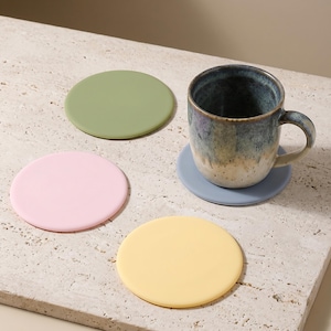May include: A set of four circular coasters in solid pastel colours including sage green, light pink, pale yellow, and soft blue. One blue coaster supports a speckled ceramic mug with a blue and beige gradient finish and a curved handle.