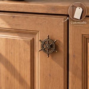 Peut inclure: Un bouton de placard en métal couleur bronze en forme de barre de navire à six rayons, fixé sur une porte de placard en bois. Un petit encart circulaire dans le coin supérieur droit affiche une étiquette blanche avec le texte Customizable.