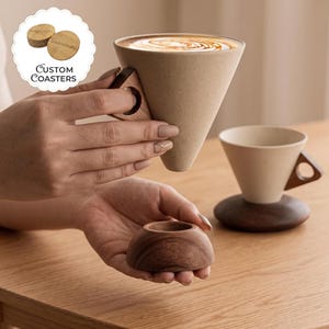 May include: A cone-shaped ceramic mug with a speckled beige finish and a triangular wooden handle. A separate dark wood circular base sits below, matching the one under a second mug. A circular label reads Custom Coasters.