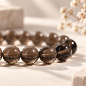 Könnte beinhalten: Ein Armband aus glatten, runden, durchscheinenden Rauchquarzperlen. Die Perlen sind dunkelbraun und haben eine polierte, reflektierende Oberfläche. Die Perlen sind in einer kreisförmigen Anordnung aufgereiht und zeigen ihre einheitliche Form.