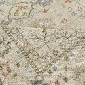 May include: Beige area rug featuring a detailed floral design in blue, green, and rust hues. The rug has a distressed appearance, with a folded section showing the underside. The word "TARIKID" is visible.
