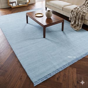 Pode incluir: Um tapete retangular azul-claro com uma superfície texturizada e detalhes de franjas nas extremidades curtas. O tapete apresenta o texto Fahad rugs impresso em branco no canto inferior direito, proporcionando uma base de cor sólida.