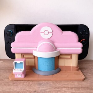 Könnte beinhalten: Ein 3D-gedrucktes Modell eines Gebäudes in Rosa und Beige mit einer blauen Tür und einem kleinen Spielautomaten. Das Modell hat ein weißes und rosa Logo auf dem Dach. Eine schwarze Nintendo Switch-Konsole befindet sich hinter dem Modell.