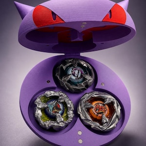 May include: A purple toy case shaped like a cartoon character's head, with red eyes and pointed ears. The case is open, revealing three colorful spinning tops inside. The tops have intricate designs and metallic accents.