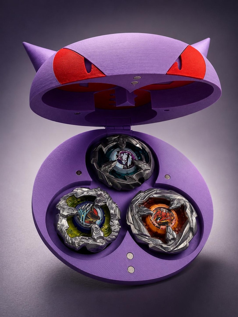 May include: A purple toy case shaped like a cartoon character's head, with red eyes and pointed ears. The case is open, revealing three colorful spinning tops inside. The tops have intricate designs and metallic accents.