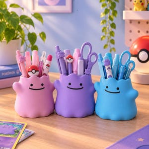 Peut inclure: Trois organisateurs de bureau en silicone en forme de Ditto de Pokemon, en rose, violet et bleu. Chaque récipient présente un simple sourire noir et deux points pour les yeux, contenant divers stylos, crayons et ciseaux avec des motifs.