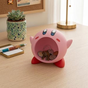 May include: A pink Kirby-shaped desk organizer with a circular opening, holding keys and coins. The organizer has red feet and small arms. The desk also features a succulent in a speckled green pot, a notepad, and a pen.