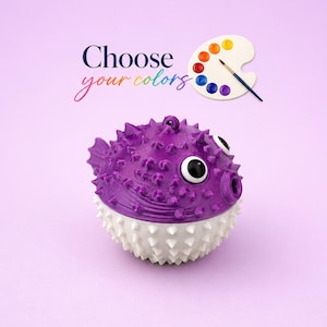 Puede incluir: Una figura de pez globo impresa en 3D con la mitad superior morada y la mitad inferior blanca, con espinas prominentes, ojos grandes en blanco y negro y un pequeño lazo en la parte superior. El texto "Choose your colors" aparece arriba con un icono de paleta.