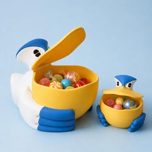 Pode incluir: Duas taças de cerâmica em forma de pelicano. O pelicano maior é branco com cabeça e pés azuis e bico amarelo. O pelicano menor é uma versão em miniatura. Ambas estão cheias de doces coloridos.