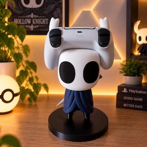 Könnte beinhalten: Eine weiß-dunkelblaue Hollow Knight-Charakterfigur, die als Controller-Ständer dient. Die Figur hat einen großen weißen Kopf mit schwarzen ovalen Augen und einen dunkelblauen Umhang und trägt einen weißen Sony PlayStation-Controller.