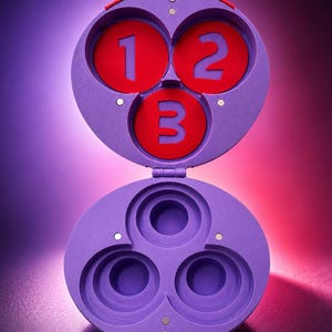May include: A purple, hinged container with three circular compartments. The top section has three red circles with the numbers 1, 2, and 3. The bottom section has three empty circular spaces. The container is open, revealing the interior.