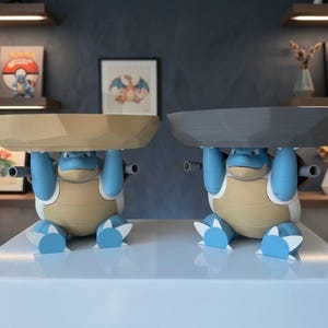 Może przedstawiać: Dwie niebiesko-beżowe figurki Blastoise, z których każda trzyma nad głową dużą, fasetowaną tacę. Jedna taca jest beżowa, a druga ciemnoszara. Figurki mają niebieskie kończyny, beżową skorupę i dwa działa wystające z tyłu.