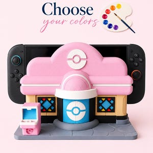 Puede incluir: Una consola Nintendo Switch rosa y negra con un soporte decorativo personalizado rosa, blanco, azul y beige. El soporte está diseñado para parecer un edificio. La imagen incluye el texto "Elige tus colores" y una paleta de pintura.