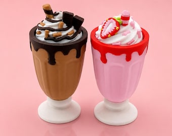 Puede incluir: Dos figuras en miniatura de batidos sobre bases blancas. La de la izquierda es marrón con sirope de chocolate negro, cubierta con nata blanca, trozos de chocolate y barquillos. La de la derecha es rosa con sirope rojo, cubierta con nata blanca y una rodaja de fresa.