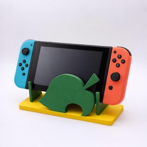 Peut inclure: Une console Nintendo Switch est posée sur un support en bois jaune et vert. Le support présente un motif de feuille verte et soutient l'écran et les manettes bleues et oranges de la console.