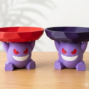 Può includere: Due figure viola del personaggio Gengar, ognuna delle quali sostiene una ciotola sfaccettata sopra la testa. Una ciotola è rosso brillante e l'altra è viola scuro. Entrambe le figure hanno occhi rossi e un ampio sorriso bianco su una superficie.