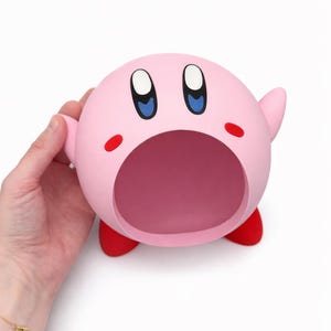 May include: A spherical pink Kirby character figure featuring a large open mouth, oval blue and black eyes, small red cheek marks, and two small red feet. The figure has a smooth matte finish and is shaped like a hollow container or bowl.