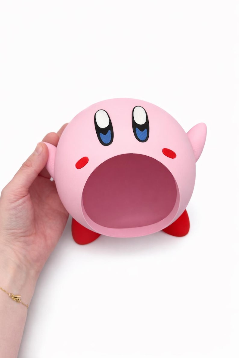 May include: A spherical pink Kirby character figure featuring a large open mouth, oval blue and black eyes, small red cheek marks, and two small red feet. The figure has a smooth matte finish and is shaped like a hollow container or bowl.