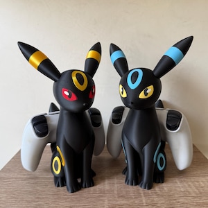 Op de afbeelding: Twee controllerstandaards geïnspireerd op Umbreon, elk met een witte gamecontroller. De linkerfiguur heeft gele ringen en rode ogen, terwijl de rechterfiguur blauwe ringen en gele ogen heeft. Beide zijn gladde, matzwarte, vosachtige figuren.