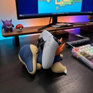 Puede incluir: Un soporte para mando con forma de Cyndaquil impreso en 3D en azul, beige y naranja, sosteniendo un mando de juego blanco y negro. Una figura de Gengar morada y una Pokéball roja y blanca están en una estantería de madera.