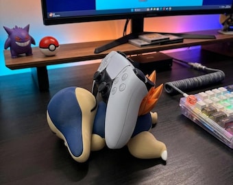 May include: A 3D-printed Cyndaquil-shaped controller holder in blue, tan, and orange, holding a white and black game controller. A purple Gengar figure and a red and white Pokeball are on a wooden shelf in the background.