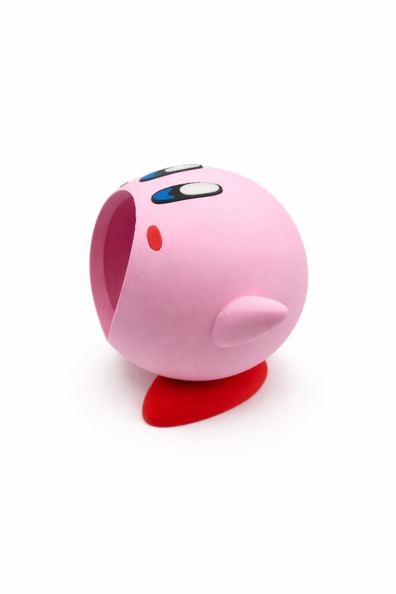 May include: A pink, spherical figurine with a red base, featuring large eyes and a small mouth. The figurine has a small, pointed ear-like protrusion on its side. The design is reminiscent of a popular video game character.