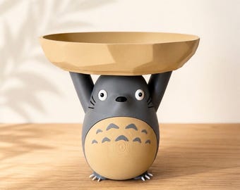 May include: A dark gray and tan decorative bowl featuring a stylized creature with large white eyes, pointed ears, and a rounded belly with chevron markings. The creature has its arms raised upward to support a wide, shallow, tan-colored bowl.