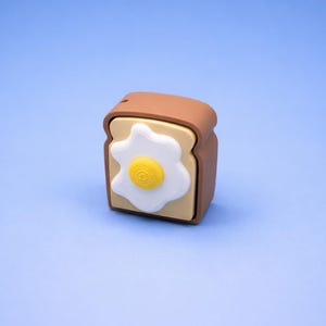 Op de afbeelding: Een bruin en beige toastvormig object met een gebakken ei-ontwerp. Het ei heeft een witte omtrek en een gele dooier. Het object staat op een lichtblauwe achtergrond.