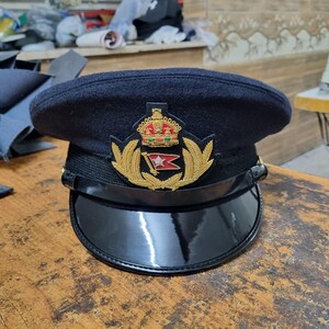 May include: A navy blue captain's hat with a black, shiny brim. The hat features a gold emblem with a crown, laurel leaves, and a red and white flag. The hat is sitting on a wooden surface.
