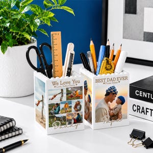 May include: Two white personalized pen holders with photos and text. One says "We Love You" and "Daddy", the other says "Best Dad Ever" and "Papa". Each holds pens, pencils, scissors, and a ruler.