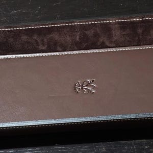 May include: A rectangular brown leather tray with a dark brown suede interior and white stitching. The tray features a silver emblem in the center. The corners are curved and the edges are black.