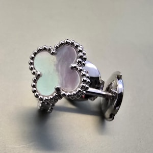 May include: A silver-toned stud earring featuring a four-leaf clover shape with a pearlescent, iridescent inlay. The clover is bordered by a beaded metal edge, and the earring includes a matching silver-toned post and a circular butterfly back.