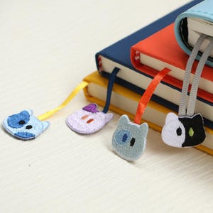 以下が含まれることがあります： 青、紫、グレー、白黒の猫の形をした刺繍のブックマーク。各ブックマークには、異なる色のリボンが付いています。ブックマークはカラフルな本に挟まれています。