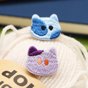 May include: Two embroidered cat-shaped patches. The top patch is light blue with dark blue spots and black eyes. The bottom patch is light purple with a dark purple ear detail and two orange eyes. Both patches feature detailed stitching.