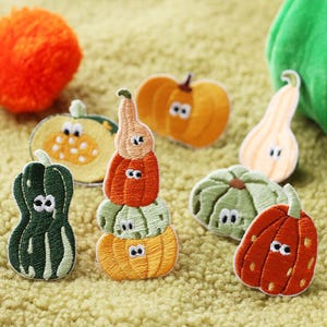 May include: A collection of embroidered pumpkin patches in shades of orange, green, and yellow. Some are stacked, all featuring embroidered eyes and the number eight. The patches are displayed on a textured, light-yellow surface.