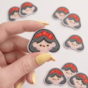 May include: A small embroidered patch featuring a stylized character with dark hair, a bright red headband with a bow, and a light skin tone. The patch has a white border and is shaped like a head. Several identical patches are scattered nearby.