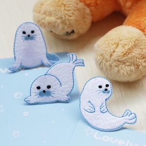 Puede incluir: Tres parches bordados que representan crías de foca blancas con detalles en azul claro. Cada parche muestra una foca en una pose diferente, incluyendo una sentada, una tumbada y una curvada, todas con ojos negros y detalles faciales cosidos.