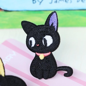 May include: Embroidered patch of a black cat with large white eyes, a pink nose, and a pink collar with a yellow charm. The cat has purple inner ears and a small tail. The patch is on a pink and white striped surface.