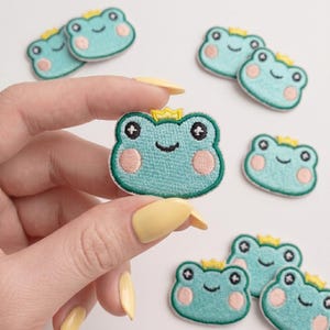 May include: A collection of embroidered iron-on patches shaped like frog faces. Each patch features a light green frog with a small yellow crown, black eyes with white highlights, and round pink cheeks. The embroidery has a textured finish.