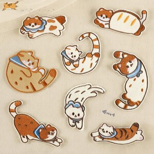 Puede incluir: Una colección de ocho parches bordados termoadhesivos que muestran varios gatos en diferentes posturas, incluyendo gatos con collares azules, un gato con forma de pan y un pequeño icono de espina de pescado, todo en tonos marrón, blanco y naranja.