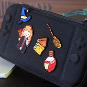 Peut inclure: Étui de transport noir orné d'écussons brodés représentant un chapeau de sorcier, une sorcière avec une baguette en forme de cœur, des livres, un balai et une bouteille de potion. L'étui a un design de console de jeu.