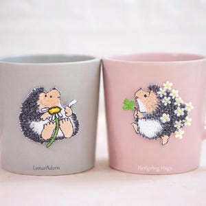 Könnte beinhalten: Ein grauer Becher mit einem gestickten Igel, der ein Gänseblümchen hält, und dem Text LunarAdorn, neben einem rosa Becher mit einem gestickten Igel, der ein vierblättriges Kleeblatt und weiße Blumen hält, mit dem Text Hedgehog Hugs.