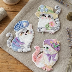 Puede incluir: Tres parches bordados de gatos. Un gato lleva una boina azul con una flor blanca. Otro gato lleva un sombrero de paja amarillo con flores azules y sostiene una flor rosa. El tercer gato lleva un gorro morado con un lazo y detalles amarillos.