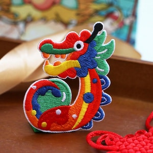May include: An embroidered patch shaped like a stylized dragon with a curved body. The design features vibrant red, orange, yellow, green, and blue thread colors. The dragon has a prominent head with a white eye, a yellow snout, and a green mane.
