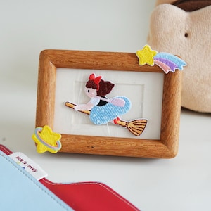 May include: A rectangular wooden frame featuring embroidered patches: a girl with a red bow flying on a broomstick in the center, a yellow shooting star with a rainbow tail in the top right, and a yellow planet with a ring in the bottom left corner.